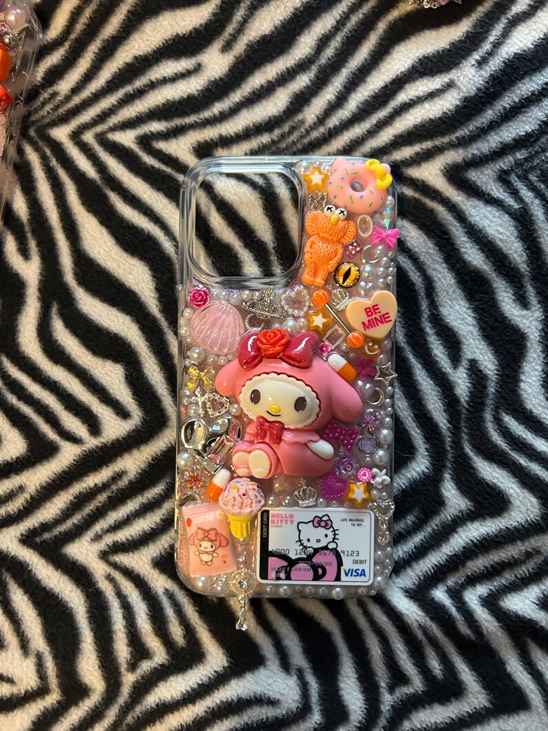 May include: Clear phone case decorated with a pink My Melody figure, a heart that says "Be Mine", and various charms. The case also features a Hello Kitty credit card design and is embellished with pearls and other decorative elements.