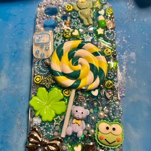 May include: A custom-made phone case with a whimsical design. The case features a large, swirled lollipop, a teddy bear, a four-leaf clover, and various charms. The background is a mix of glitter and beads, with a silver bow and a small phone charm.