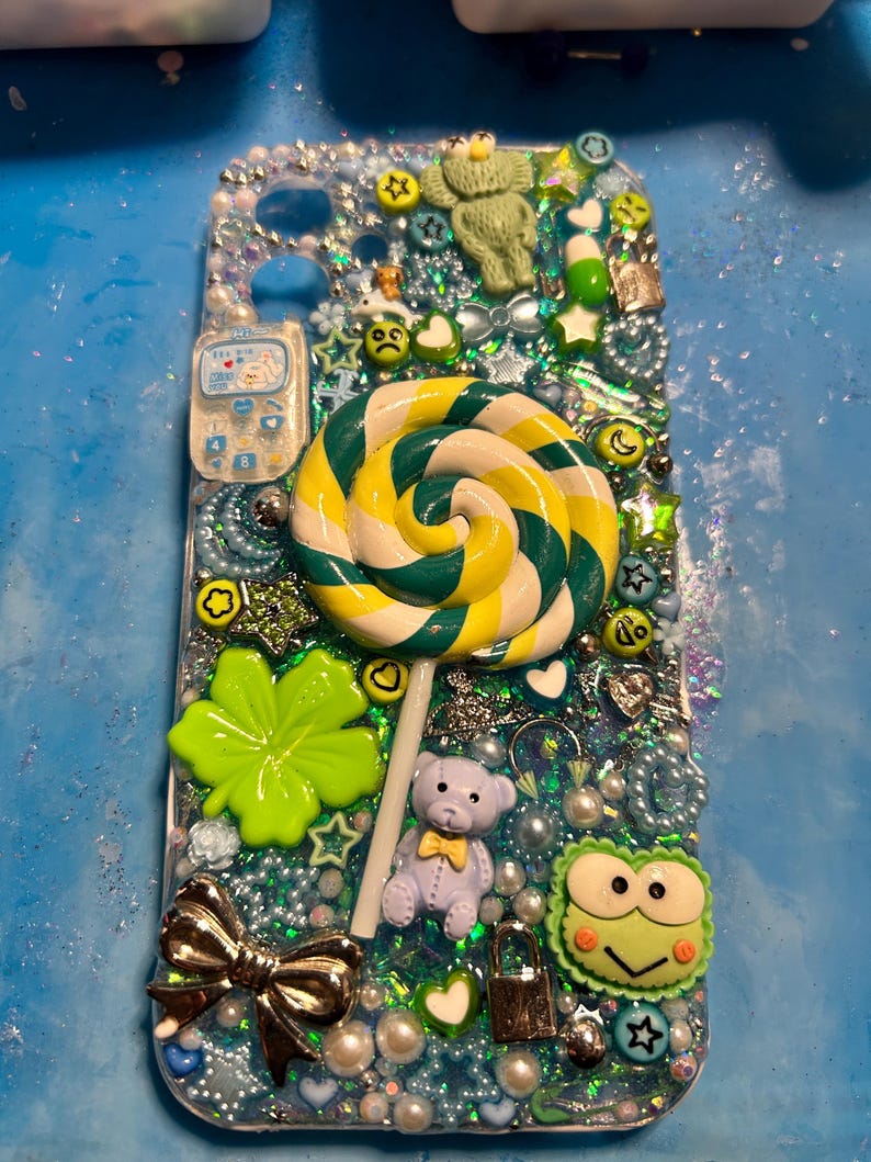 May include: A custom-made phone case with a whimsical design. The case features a large, swirled lollipop, a teddy bear, a four-leaf clover, and various charms. The background is a mix of glitter and beads, with a silver bow and a small phone charm.
