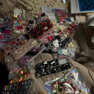 May include: A collection of decorative phone cases, featuring various designs with rhinestones, beads, and charms. The cases showcase a range of colors, including pink, black, and clear, with some cases featuring cartoon characters and floral patterns.