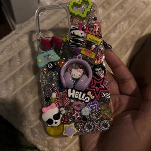 May include: Clear phone case decorated with various charms, including a Hello Kitty design with headphones, a skull with a pink bow, a green heart, and a Haribo candy package. The word "Hello" is also visible.
