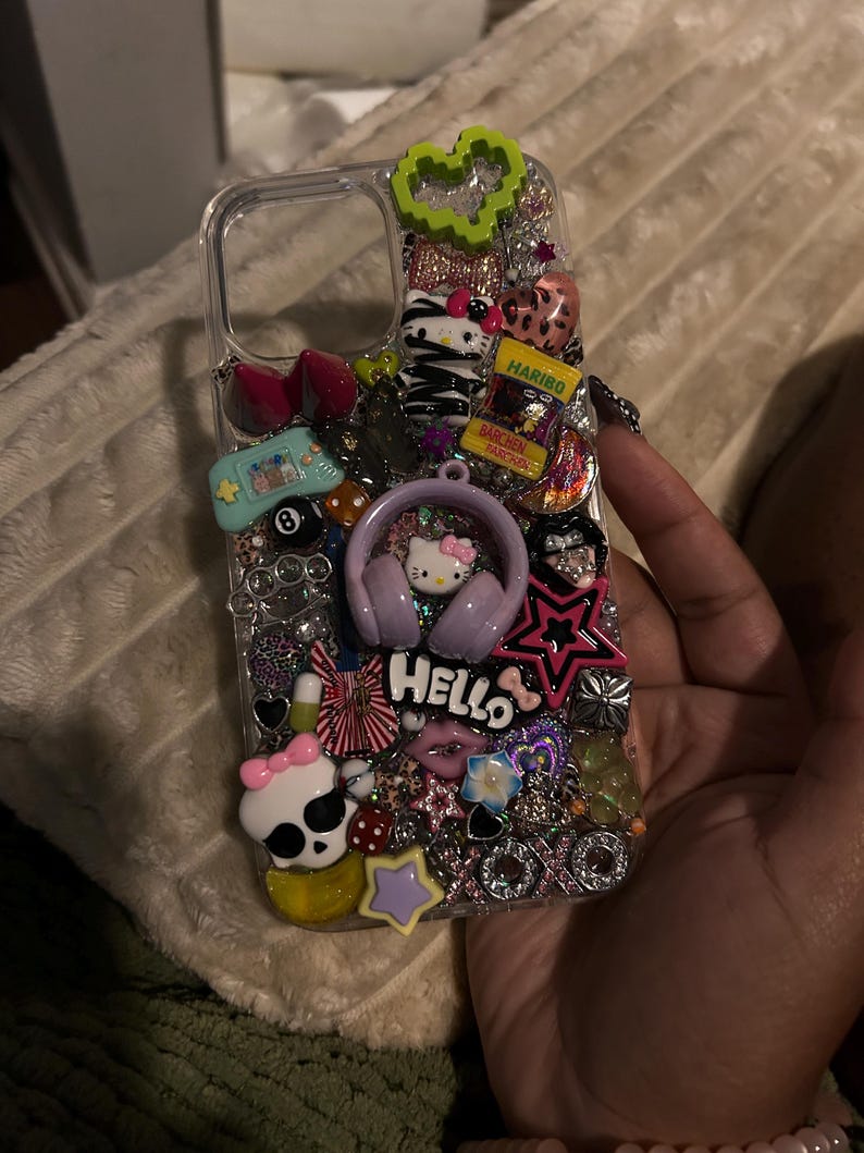 May include: Clear phone case decorated with various charms, including a Hello Kitty design with headphones, a skull with a pink bow, a green heart, and a Haribo candy package. The word "Hello" is also visible.