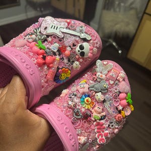 May include: Pink Crocs decorated with colorful charms, including a guitar, a skull, a rainbow, and a variety of cartoon characters. The charms are attached to the shoes with rhinestones and glitter.