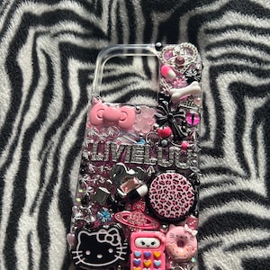 Könnte beinhalten: Eine transparente Handyhülle mit einem pink-schwarzen Design. Die Hülle ist mit Strasssteinen, Glitzer und verschiedenen Charms verziert, darunter ein Hello-Kitty-Charakter, ein Donut, ein Taschenrechner und ein Leopardenmuster. Der Text "LIVE LOUD" ist auf der Hülle sichtbar.