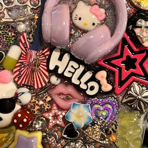 May include: A decorative phone case with a variety of colorful charms. Features include a Hello Kitty, pink headphones, a star, lips, and the word "Hello". The case is covered in glitter and other embellishments.
