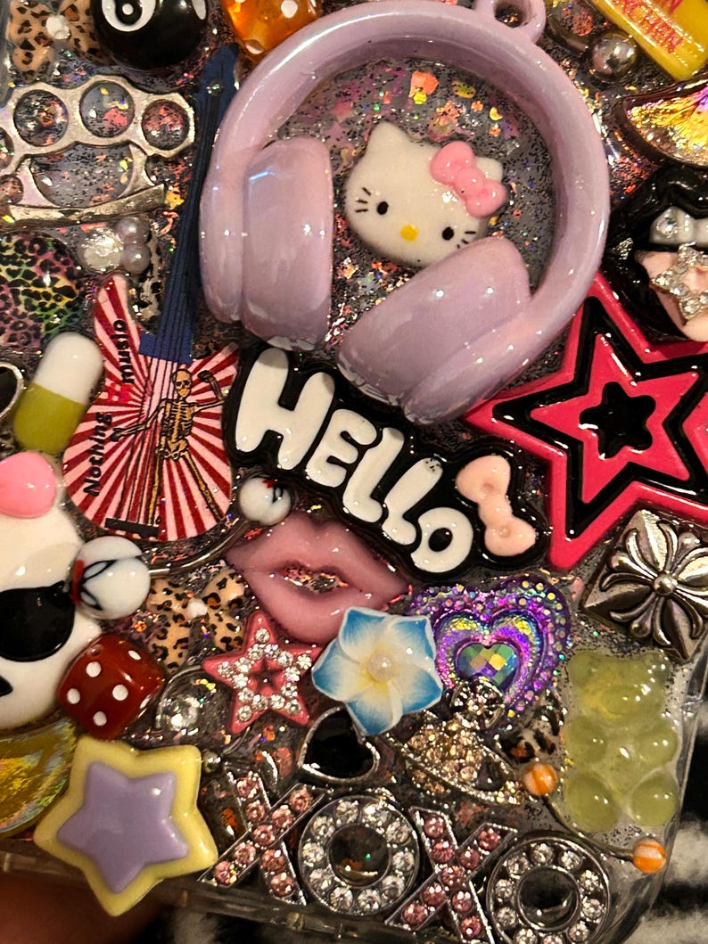 May include: A decorative phone case with a variety of colorful charms. Features include a Hello Kitty, pink headphones, a star, lips, and the word "Hello". The case is covered in glitter and other embellishments.