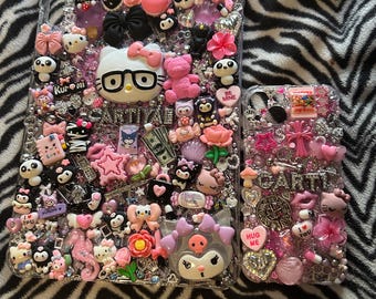 May include: Two phone cases decorated with a variety of colorful charms, including Hello Kitty, a skull, flowers, and hearts. The cases are clear and show the phone screen through the back. The cases are on a black and white zebra print background.