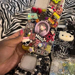 May include: Clear phone cases decorated with various charms, including a Hello Kitty design, a green pixelated heart, and a Haribo candy package. The cases are embellished with rhinestones and other decorative elements.