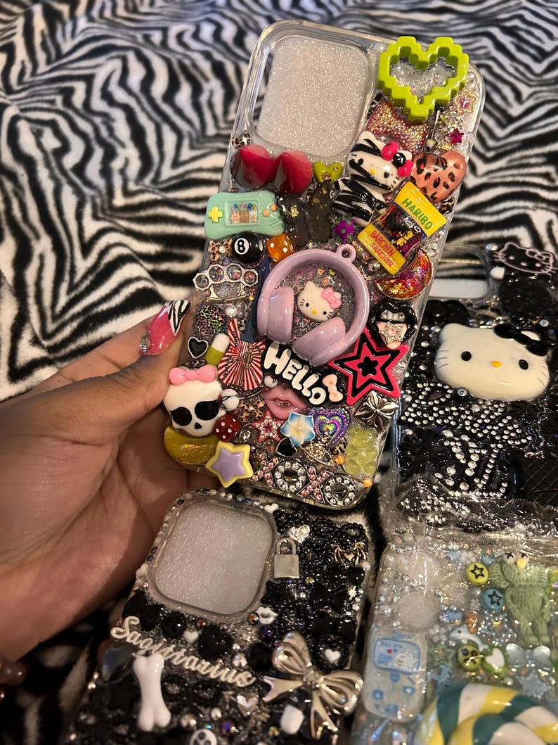 May include: Clear phone cases decorated with various charms, including a Hello Kitty design, a green pixelated heart, and a Haribo candy package. The cases are embellished with rhinestones and other decorative elements.