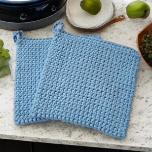 May include: Two light blue, square crochet potholders with loop handles. The potholders are made of a tightly woven, textured yarn. The potholders are sitting on a white speckled countertop.