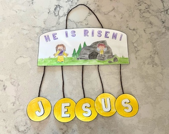 Puede incluir: Una manualidad de papel amarilla con las palabras "¡Ha resucitado!" y un dibujo de Jesús y un ángel. La palabra "JESÚS" está escrita en cinco círculos amarillos que cuelgan de la manualidad.