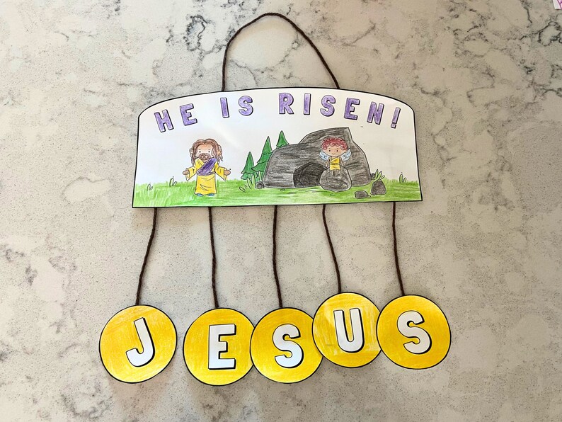 Puede incluir: Una manualidad de papel amarilla con las palabras "&iexcl;Ha resucitado!" y un dibujo de Jes&uacute;s y un &aacute;ngel. La palabra "JES&Uacute;S" est&aacute; escrita en cinco c&iacute;rculos amarillos que cuelgan de la manualidad.