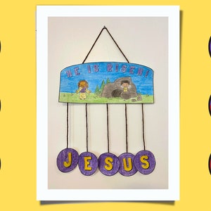 Puede incluir: Una manualidad de papel morado y azul con las palabras "He is Risen!" y un dibujo de una persona saliendo de una cueva. La manualidad tiene cinco c&iacute;rculos morados colgando de ella con la palabra "JESUS" escrita en letras amarillas.