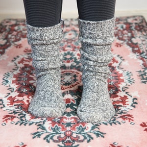 May include: A pair of gray and white speckled knit socks, bunched around the ankles, are visible. The socks are worn over dark gray leggings. The socks are on a pink rug with a floral pattern in red, green, and white.