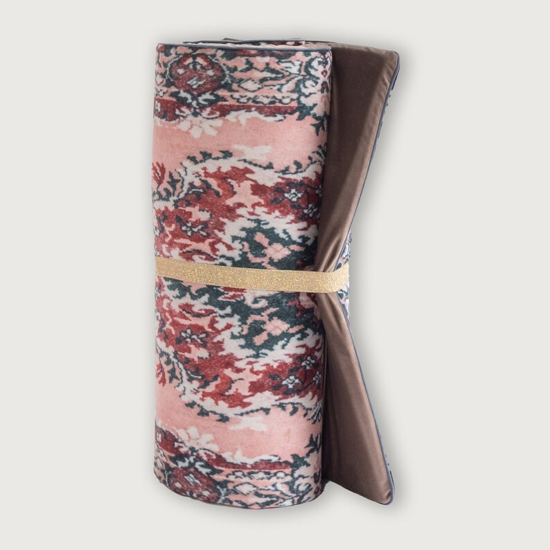 May include: Rolled-up rug with a floral pattern in shades of pink, red, and green. The rug is secured with a gold elastic band. The edges of the rug are a dark brown color.