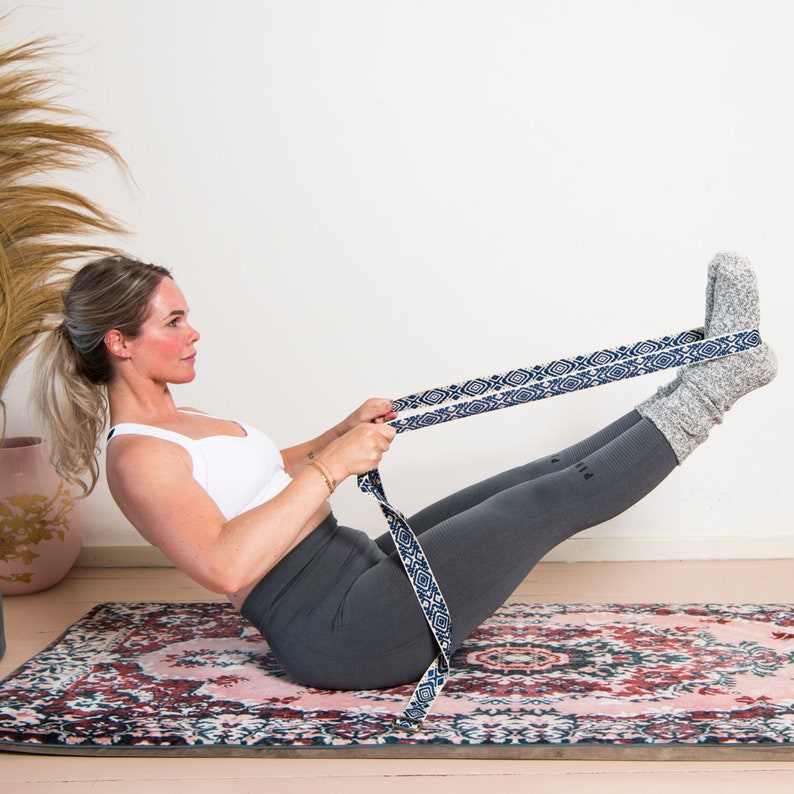 May include: A woman is doing yoga on a patterned rug, using a blue and white patterned yoga strap. She is wearing gray leggings, a white sports bra, and gray socks. The yoga strap is stretched between her hands and feet.