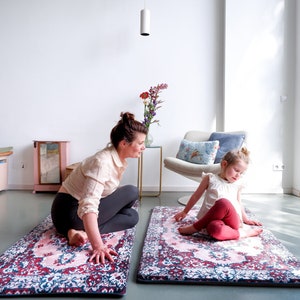 May include: Two yoga mats with a floral, Persian rug design in pink, red, and blue. A woman and a child are sitting on the mats in a bright room. The mats have a dark blue border. The woman is wearing a light-colored shirt and gray leggings.