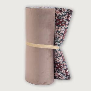 May include: Rolled-up area rug with a muted pink velvet exterior and a patterned interior. The rug is secured with a gold ribbon. The patterned section features a design with red, white, and green accents.