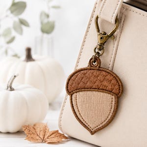 May include: An acorn-shaped bag charm featuring a quilted brown cap and a light beige textured body. The charm is attached to a brass-toned swivel lobster clasp and is currently clipped onto the side of a cream-colored handbag.
