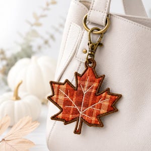 May include: A fabric maple leaf keychain featuring an orange and brown plaid pattern with stitched white veins. The leaf is attached to a brass-toned metal lobster clasp, which is clipped onto the handle of a white textured handbag.