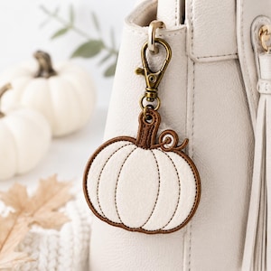 May include: A pumpkin-shaped keychain featuring a white fabric center with stitched vertical lines and a brown fabric border. The pumpkin has a small brown curly vine detail at the top and is attached to a bronze-colored metal lobster clasp.