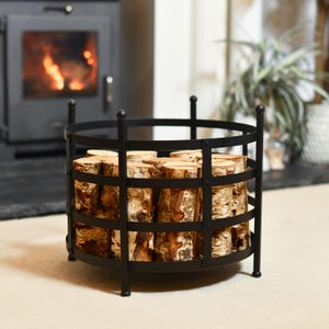 May include: Black metal log holder with a round base and a grid design. The holder is filled with stacked logs of wood.