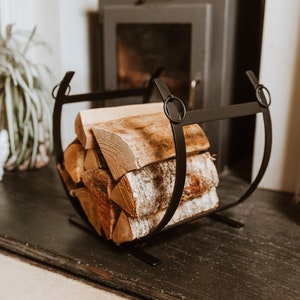 May include: Black metal log holder with a stack of firewood inside. The holder has two rings on the sides and is sitting in front of a fireplace.