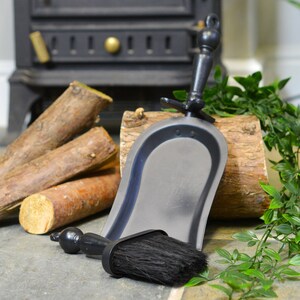 K&ouml;nnte beinhalten: Ein schwarzes Kamin-Set mit Kehrschaufel und B&uuml;rste, mit Holzgriffen und schwarzen Borsten. Das Set steht auf einer Steinoberfl&auml;che neben einem Stapel Brennholz und einem Holzscheit.