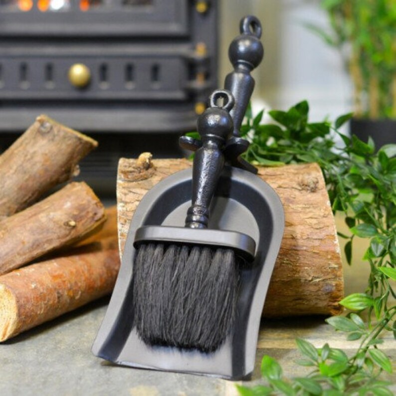 K&ouml;nnte beinhalten: Ein schwarzes Kaminb&uuml;rsten- und Kehrblech-Set. Die B&uuml;rste hat einen dunklen Borstenkopf und ist an einem dekorativen Griff befestigt. Das Kehrblech hat eine passende schwarze Farbe. Das Set wird auf einem Holzst&uuml;ck mit Brennholz und Gr&uuml;nzeug im Hintergrund pr&auml;sentiert.