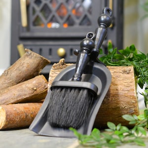 K&ouml;nnte beinhalten: Ein schwarzes Kaminb&uuml;rsten- und Kehrblech-Set. Die B&uuml;rste hat einen dunklen Borstenkopf und ist an einem dekorativen Griff befestigt. Das Kehrblech hat eine passende schwarze Farbe. Das Set wird mit Brennholz und Gr&uuml;nzeug pr&auml;sentiert.