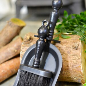 K&ouml;nnte beinhalten: Ein schwarzes Kaminb&uuml;rsten- und Kehrblech-Set. Die B&uuml;rste hat einen dunklen Borstenkopf und ist an einem dekorativen Griff befestigt. Das Kehrblech hat eine passende schwarze Farbe. Das Set wird auf einem Holzst&uuml;ck pr&auml;sentiert.