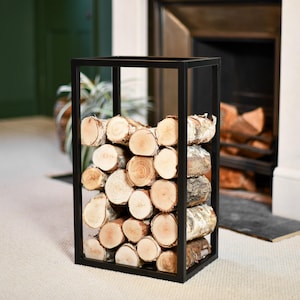 May include: A black metal log holder filled with stacked firewood. The logs are a mix of light brown and tan, with visible wood grain. The holder is rectangular and sits on a light-coloured carpet, with a fireplace in the background.