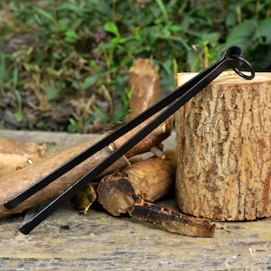 May include: Black metal fireplace tongs with a ring handle. The tongs are resting on a pile of wood logs.