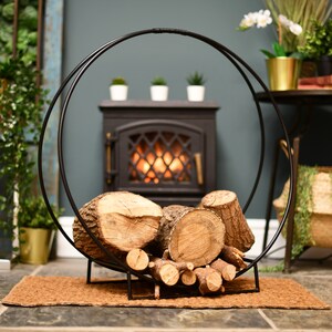 May include: Black metal log rack with a circular design, holding a stack of firewood logs in front of a fireplace.