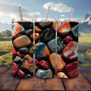 May include: Three stainless steel tumblers with a colorful abstract design of swirling orange, red, blue, and green stones. The tumblers are on a wooden surface.