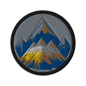May include: A round embroidered patch with a grey background and a brown, blue and yellow mountain design.