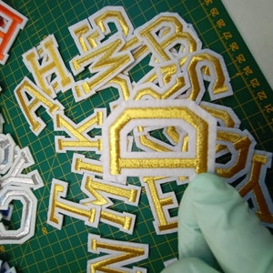 May include: A pile of gold and white felt letters, including the letters A, B, D, E, H, I, K, M, N, P, S, T, and W. The letters are arranged in a random order and are all the same size.