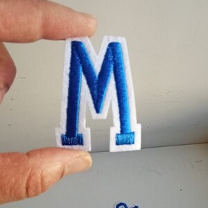May include: A white felt letter "M" with a blue embroidered outline and fill, held by a hand. Other blue and white embroidered letters are scattered below. The letters are likely for crafts or personalisation.