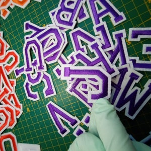 May include: Purple felt letters with white outlines on a green cutting mat. The letters are of various sizes and are arranged in a random pattern.