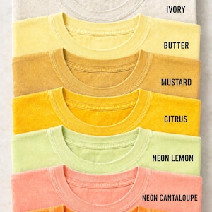May include: A stack of seven crew neck t-shirts in various colours labelled BLANK/CUSTOM PRINT at the top. The colours from top to bottom are ivory, butter, mustard, citrus, neon lemon, neon cantaloupe, and bright orange, each with a matching label.