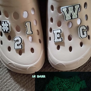 May include: Two beige clogs with white and black letter and number charms. The charms glow green in the dark. The charms spell out "#1 W.E.G. 2"