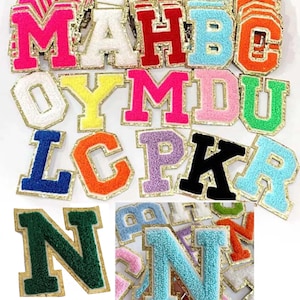 May include: A selection of colourful, embroidered varsity letter patches. The letters are in various colours, including pink, red, blue, and orange, with a gold border. The patches are made of a fuzzy material.