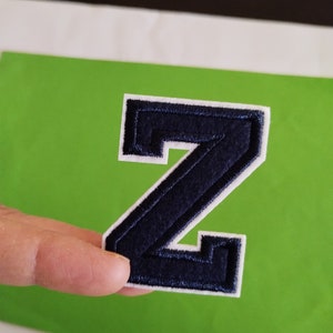 May include: A navy blue felt letter Z with white stitching around the edges. The letter is on a green background.