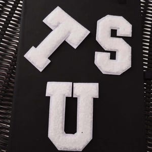 May include: Three white felt letters, "T", "S", and "U", are arranged on a black surface. The letters are in a block font style.
