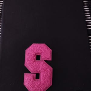 May include: A pink felt letter S, with a slightly raised, textured surface. The letter is on a black background.