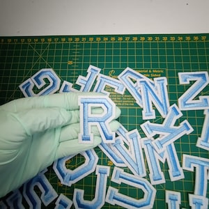 May include: A collection of light blue felt letters with white outlines. The letters are arranged in a pile on a green cutting mat. The letters are all uppercase and include the letters J, R, N, Z, and others.