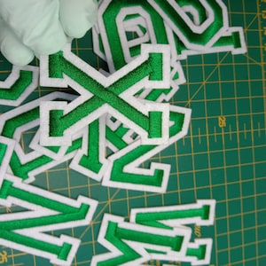 May include: Green felt letter patches with white outlines. The letters are X, N, and W. The patches are on a green cutting mat.