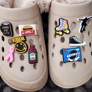 May include: A pair of beige clogs with various colourful shoe charms. The charms include a coffee cup with the text "Coffee for Life", a pink ribbon, a yellow smiley face with the text "Nirvana", a red bottle with the text "Victoria Bitter", a pink and yellow roller skate, a black and white silhouette of a person skateboarding, a black and white sneaker, a blue and white Oreo cookie package, and a red and white package with the text "Four'N Twenty Classic Meat Pie".