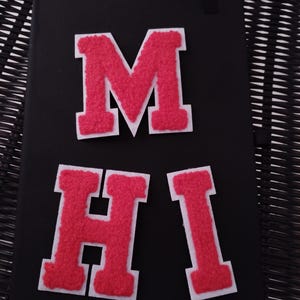 May include: Three felt letters, M, H, and I, in a bright pink colour with white outlines, are arranged on a black surface. The letters are in a varsity style font.