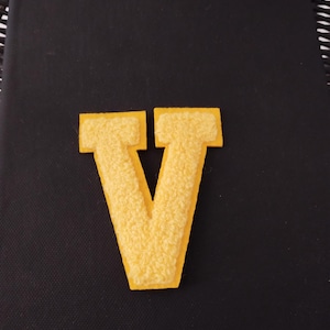 May include: A yellow felt letter V. The letter is on a black background.